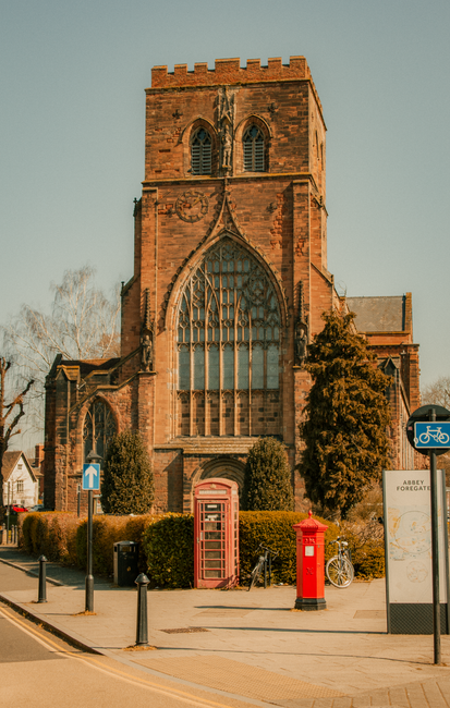 A historic red-brick church with large arched stained-glass windows and a crenellated tower, situated on a street corner with a pavement in the foreground. The scene features a traditional red British