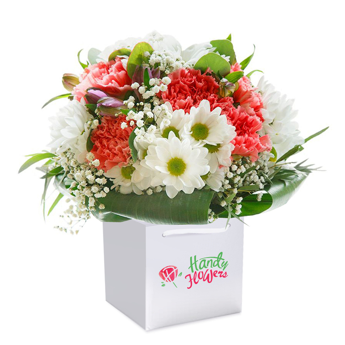 Lovingly crafted by local Basildon floral experts, this striking bouquet presents an exuberant tapestry of colour and texture, nestled upright in a contemporary white square gift bag with silky ribbon handles. The arrangement is anchored by richly hued coral and peach carnations, their ruffled, plush petals creating a sense of overflowing warmth and affection- reminiscent of the vibrant plantings at Wat Tyler Country Park. Bright, daisy-like chrysanthemums punctuate the arrangement with flashes of white and lime-green centers, introducing a spark of freshness much like morning light through the open spaces of Basildon Festival Leisure Park. Wisps of clouds-delicate gypsophila or baby's breath-float airily between the larger blooms, imbuing the bouquet with lightness and ethereal charm. On the left, a handful of purple-tinged buds, possibly alstroemeria, remain tightly closed, offering a hint of mystery and symbolising both hope and anticipation. Lush, deep green foliage frames the composition, their broad, shiny leaves wrapping the base and echoing the natural beauty of Basildon's cherished green corners. Illuminated with soft, even light, this bouquet forms a harmonious, full shape where each stem appears expertly selected and placed for maximal impact and balance. The clean lines of the gift bag and discreet ribbon handles ensure easy, stylish delivery, making this bouquet perfect for marking anniversaries, birthdays, heartfelt congratulations, or thank yous-delivered throughout Basildon with prompt care and attention.