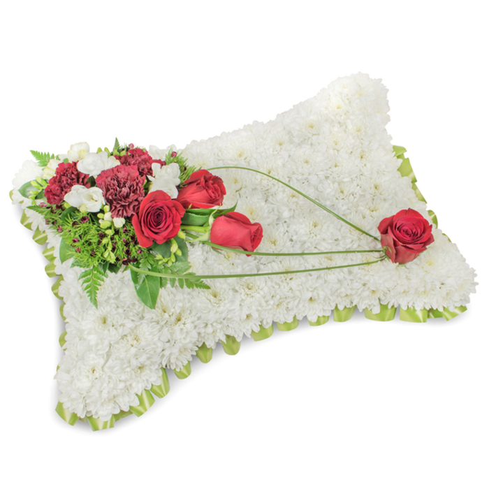 An elegant bereavement cushion, photographed with care against a neutral white setting, presents itself as a rectangular pillow completely covered with hundreds of compact, pure-white chrysanthemum heads that form a sumptuous, fleece-like surface. Along the periphery a narrow, pale-green trim-either satin ribbon or delicate foliage-peeks through the flowers, giving the piece a neat, finished outline. Nestled into the upper-left corner is an abundant cluster of deep red blooms: several velvety roses in varying openness and rich, ruffled carnations in a complementary burgundy, their strong hues creating a warm, reverent contrast to the white base. Small white accent flowers, probably freesias or spray chrysanthemums, scatter light across the arrangement and soften the transition between the bold reds and the white ground. A variety of greenery-rounded leaves, ferny fronds and glossy stems-forms a verdant backdrop, supporting the reds and reinforcing the formal composition. From the main cluster a trio of slender, bright-green bear-grass stems or decorative blades sweep diagonally toward the bottom right, tracing an elegant arc and seeming to cradle three additional red roses that appear to float across the chrysanthemum field; a single red rose anchors the final point with quiet dignity. The atmosphere is hushed and contemplative, with a faint, classic floral scent and gentle studio light that accentuates the soft textures. This timeless sympathy pillow is the kind of arrangement families in Basildon-perhaps arranging a service near Festival Leisure Park or Laindon-choose when they want a clear, heartfelt message of respect and lasting remembrance.